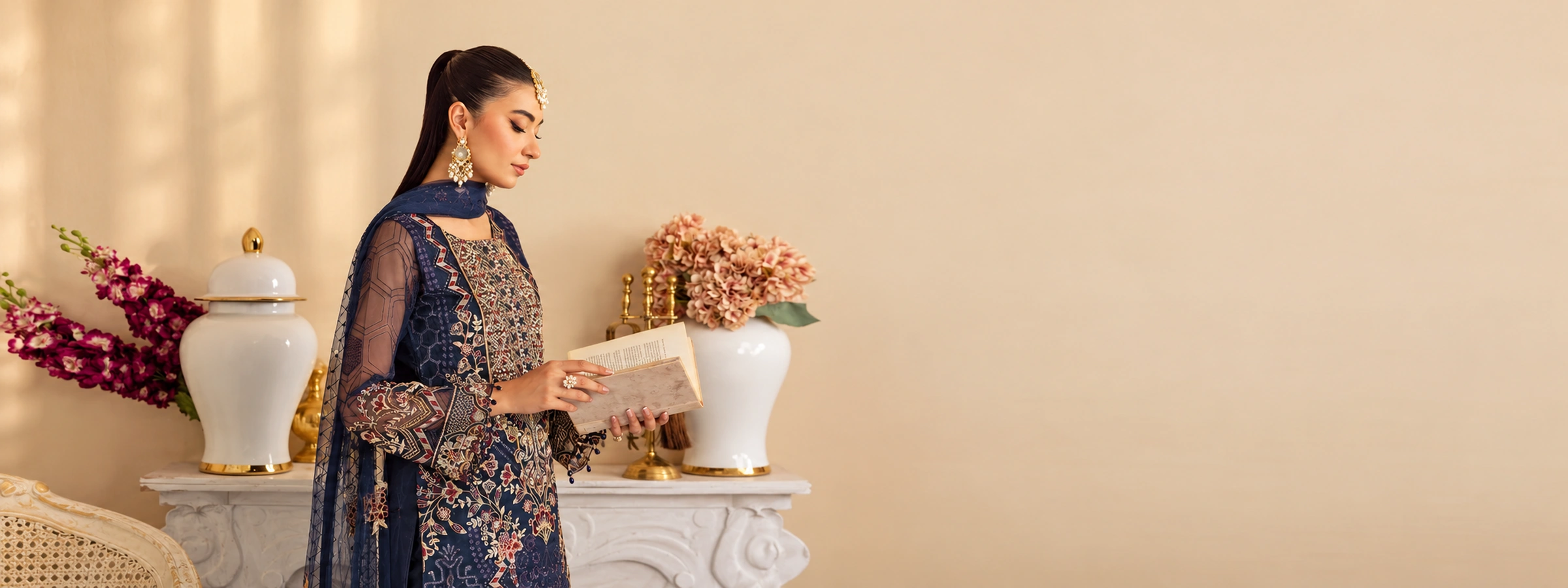 Woman in navy embroidered Pakistani suit reading a book against a cream architectural wall